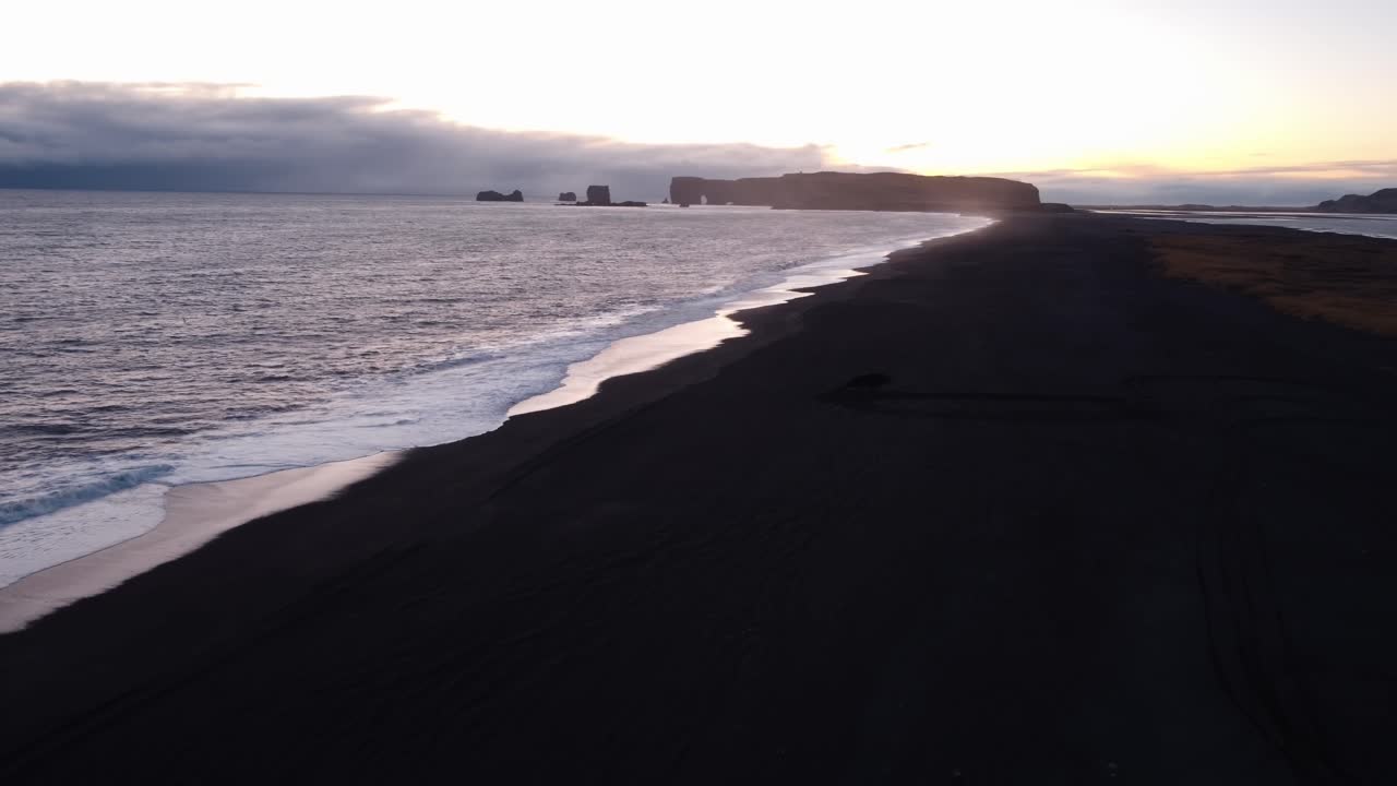 A tranquil black sand beach at Reynisfjara, Iceland, stretches into the distance, reflecting the soft hues of a sunset or sunrise over the calm North Atlantic waters