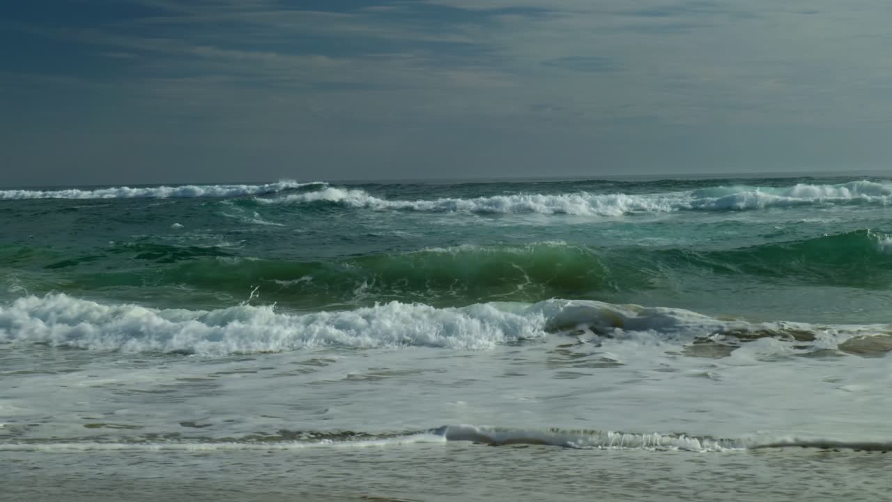 Waves break powerfully in the distance, creating dynamic whitewater as they crash, showcasing the energy of the ocean beyond the shoreline.