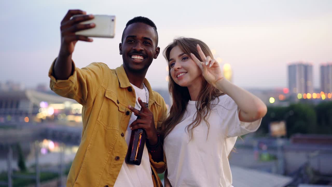 Un'attraente coppia multirazziale si sta scattando un selfie con lo smartphone a una festa sul tetto; l'uomo e la donna tengono in mano dei drink e posano con facce buffe, con la città sullo sfondo