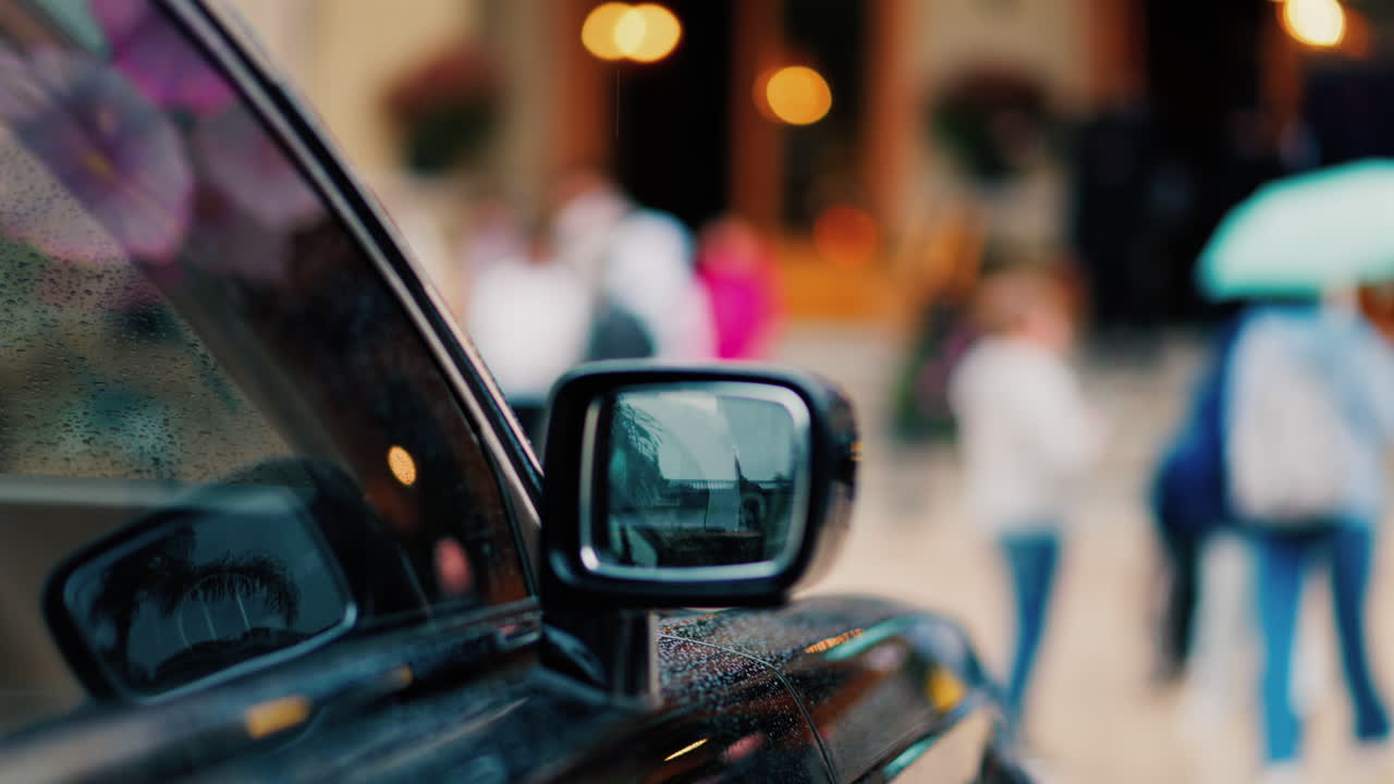 Close up of the mirror of a black car in the rain with people walking on a blurred background
