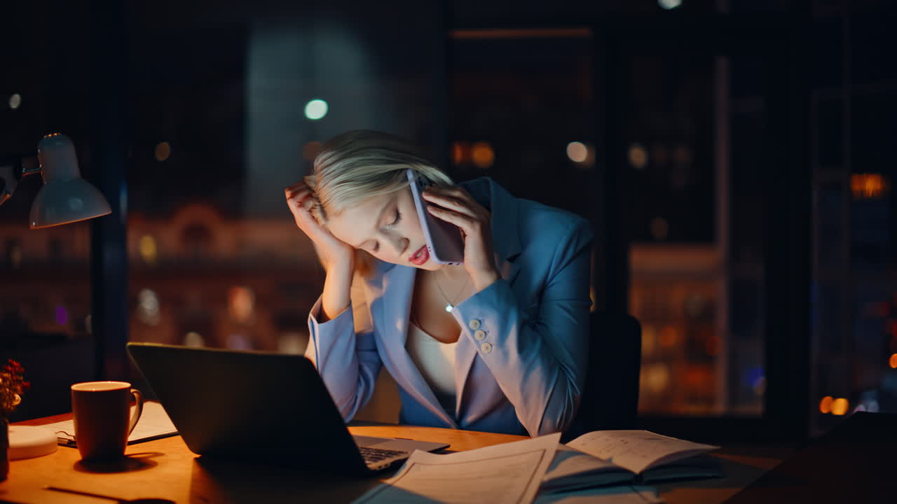 Overworked blonde talking cellphone searching documents computer table closeup