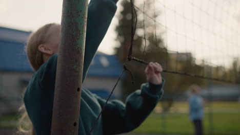 close up of girl in green sweater holding volleyball net, with blurred action of someone playing volleyball in the background, outdoor sports field with clear sky and green surroundings