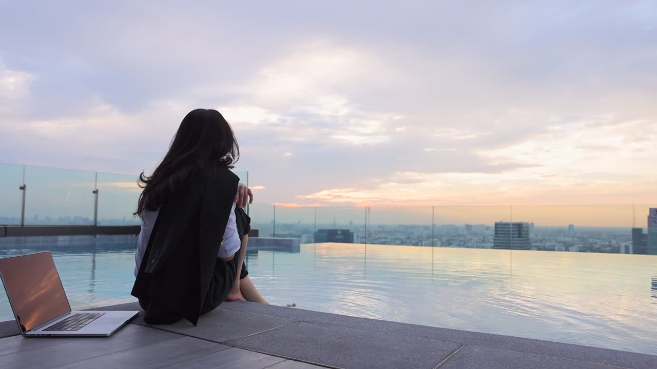 여름 여행에서 무한 수영장에서 휴식을 취하는 럭셔리 휴양 여성 아시아 소녀 관광객은 노트북에서 열심히 일한 후 도시에서 아름다운 일몰을 바라보며 휴식을 취합니다.