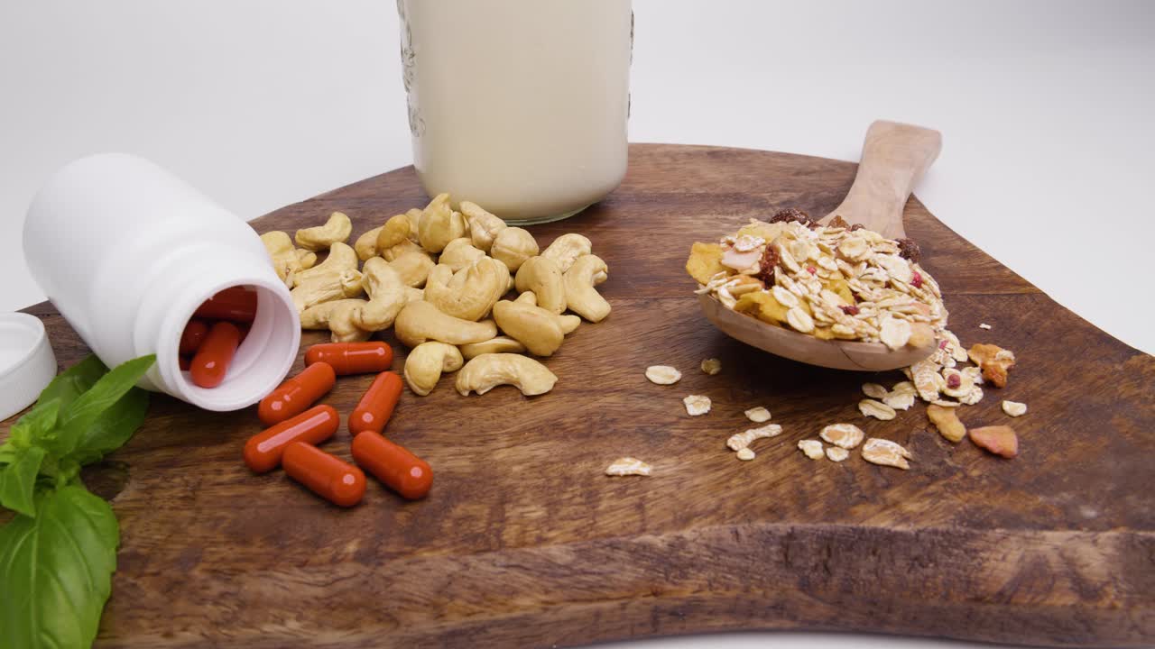 Pan shot of wooden board with supplements, cashew nuts, wooden spoon with cereals and glass filled with vegan milk on white background