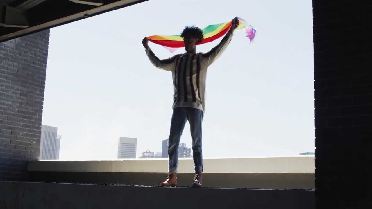 hombre afroamericano sosteniendo la bandera de la lbgt mientras estaba de pie cerca de la ventana