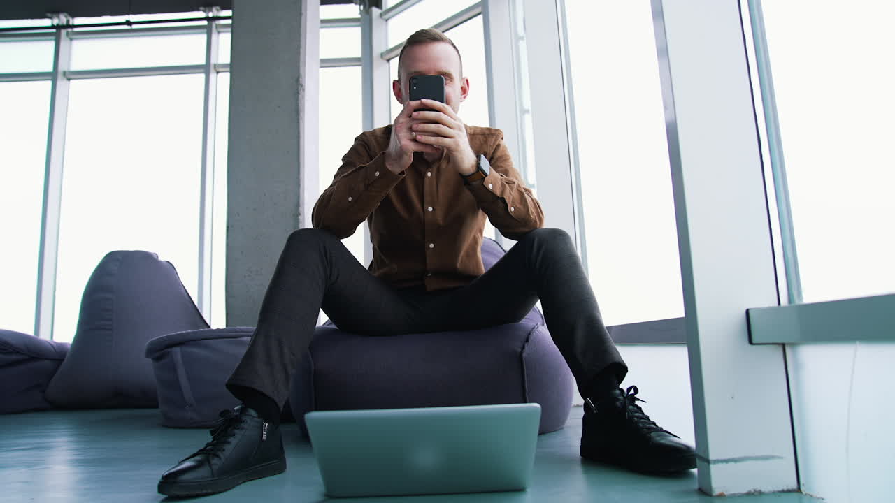 Young man with a phone in office. Handsome businessman sitting on a comfortable armchair and using a mobile phone on window background. Front view.