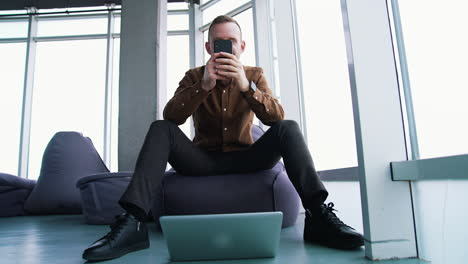 young man with a phone in office. handsome businessman sitting on a comfortable armchair and using a mobile phone on window background. front view.