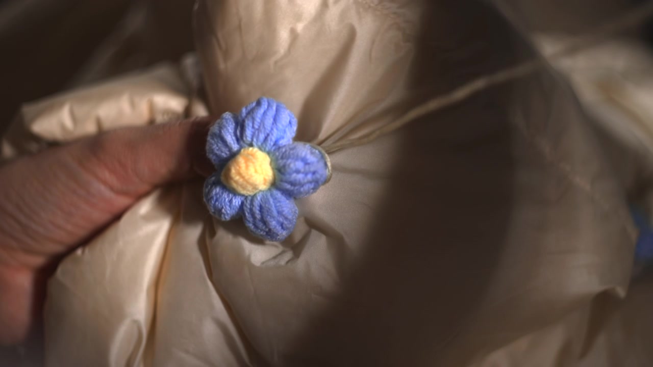 Mani in primo piano che ricamano un fiore blu su tessuto di cappotto, ricamo preciso e tensione delicata, ritocchi finali