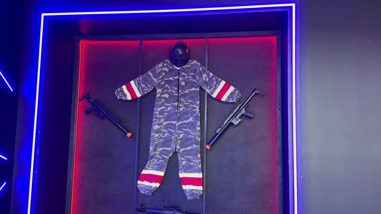Static shot of a tactical camouflage jumpsuit, black helmet, and toy guns displayed on a metal mesh wall. Vibrant blue and red neon lights illuminate this indoor laser tag or airsoft arena equipment.