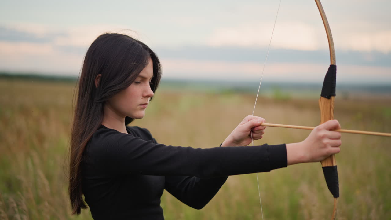 Arciera caucasica che si esercita in un campo, impugnatura in pelle, che tira e rilascia la freccia con calma e concentrazione. Le inquadrature catturano la postura di allenamento, la correzione della postura, la mentalità concentrata dell'atleta e la precisione.