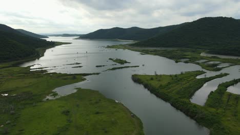 Aerial-footage-sliding-to-the-right,-taken-from-high-up,-revealing-the-gorgeous-lake-and-mountains-around-it,-a-river-feeding,-grasslands,-vehicles,-rainclouds,-provincial-landscape-in-Thailand