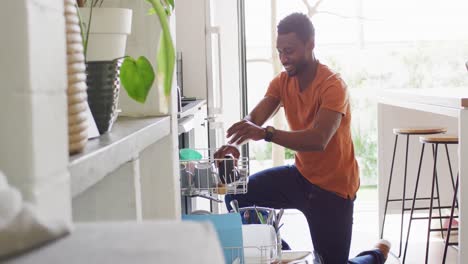 happy african american man standing in kitchen and loading dishwasher