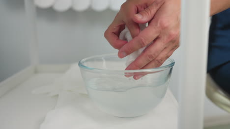 hand view of beauty expert dipping cotton towel into glass bowl of purified water on spa trolley next to lamp and cream bottles during facial treatment session in bright spa room with linen on table