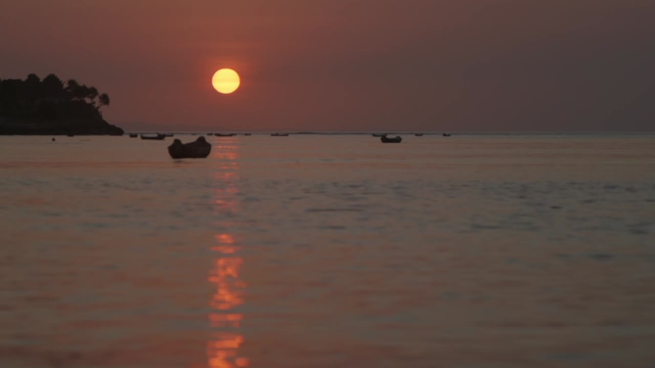 Sunset and reflection across water, Looking out past fishing boats silhouetted, between Ceningan and Lembongan Islands off Bali, high horizon sun left
