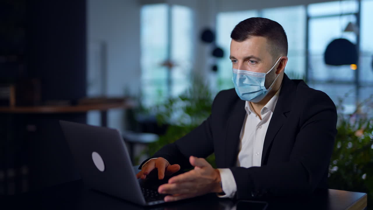 Businessman in suit and mask sitting at desk and working on computer. Entrepreneur searching information at laptop in office. Blurred background.