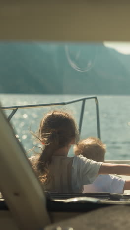 little children with outstretched arms lean out of cabin exit of motorboat in scenic sea. cheerful brother and sister sail yacht in ocean on summer holiday