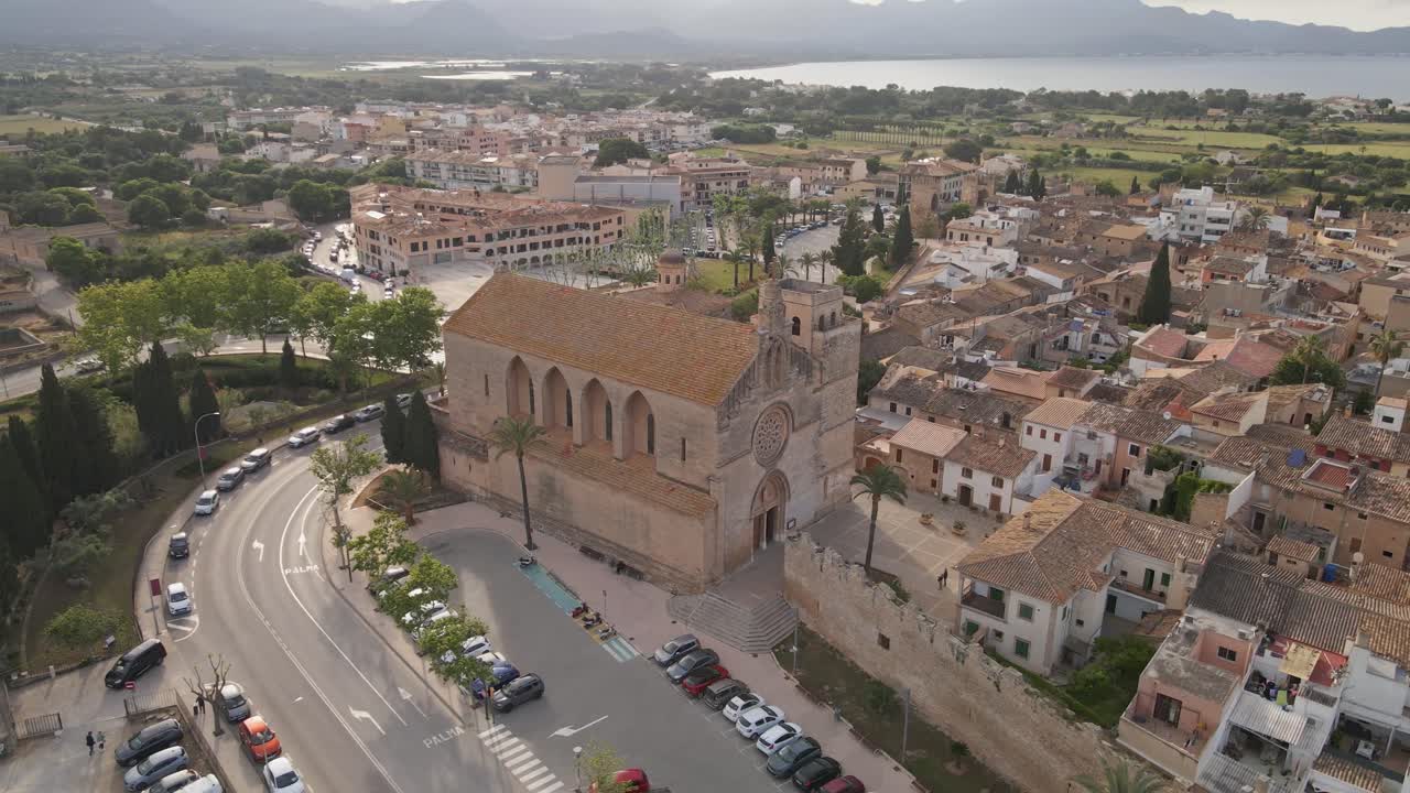 Aerial view of a Spanish historic stone church with a rose window and cross, surrounded by charming terracotta-roofed buildings in a Mediterranean village.