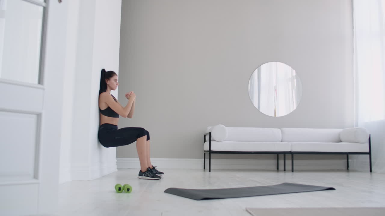 The brunette girl froze in a squat leaning on the wall doing a static exercise in a bright interior. Keep 90 degree leg angle