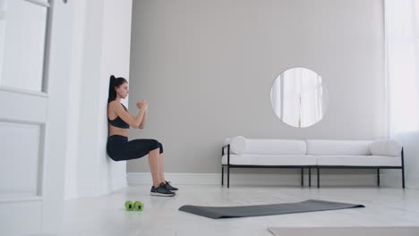 la chica morena se congeló en una cuclilla apoyándose en la pared haciendo un ejercicio estático en un interior brillante. mantener un ángulo de piernas de 90 grados