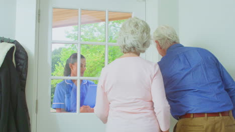 senior couple greeting female nurse or care worker making home visit in uniform at door