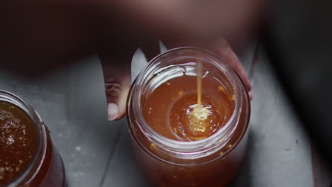vista de cerca de la mano que sostiene un frasco de vidrio, vierta y llénelo con miel de abeja extraída de líquido fresco dorado, producción apícola tradicional y apicultura en la granja de abejas