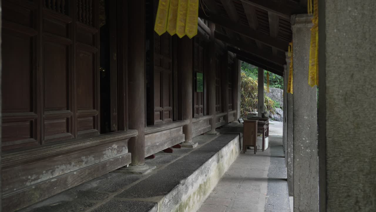 An empty wooden corridor at Bich Dong Pagoda in Ninh Binh leads past carved pillars toward soft greenery, evoking quiet solitude and temple calm, slow reveal handheld shot