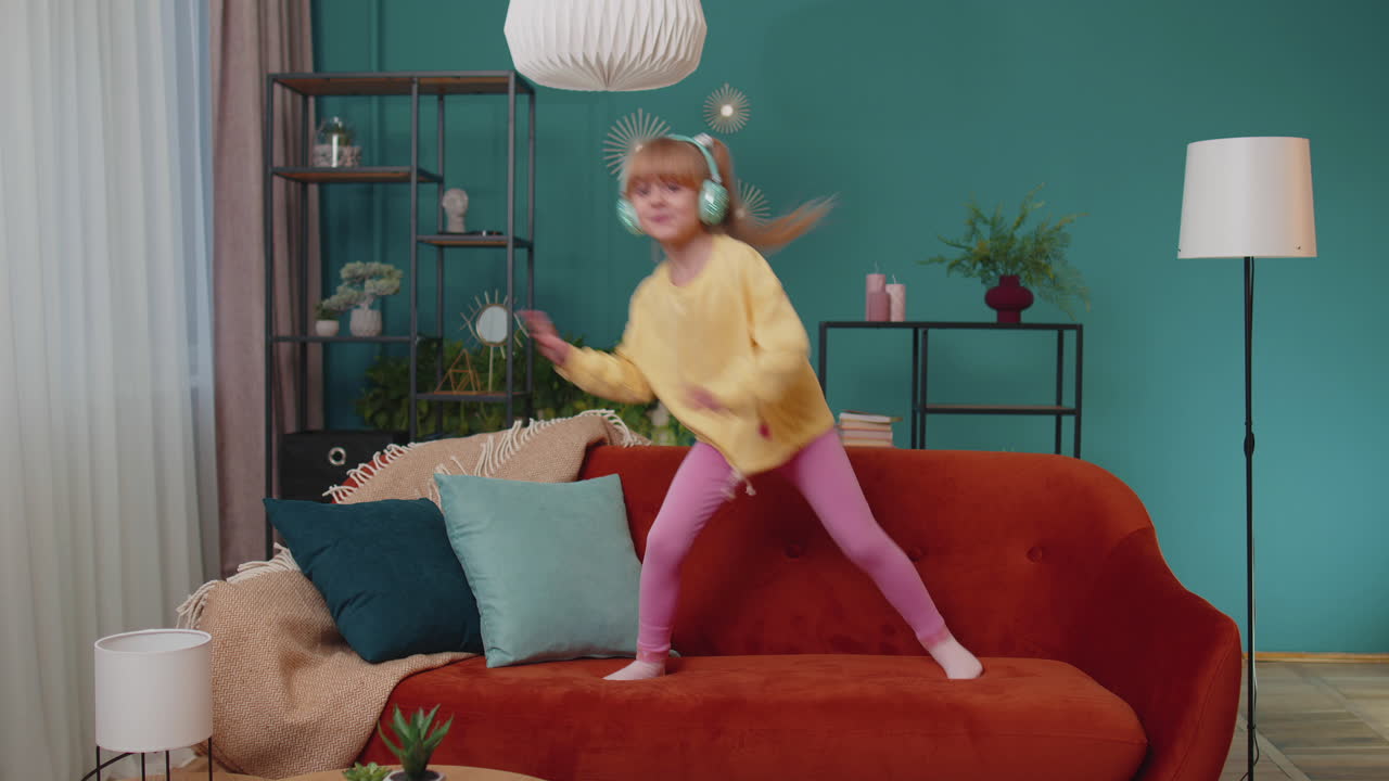 Happy child girl kid in headphones dancing and jumping while listening to the music at home alone