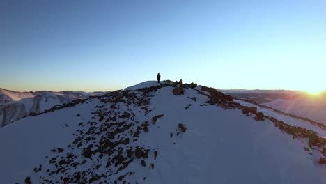 vista aérea de un montañero solitario en la cima de un pico rocoso al amanecer, nieve y sol naciente sobre las montañas, tiro de drones en órbita