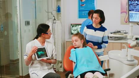 children sitting on stomatological chair listening pediatric doctor looking on tablet