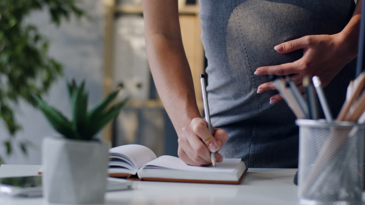 Close Up View Of The Hand Of Pregnant Businesswoman Writing Something In The Notebook With A Pen In The Office