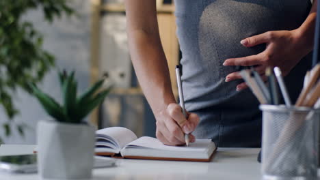 close up view of the hand of pregnant businesswoman writing something in the notebook with a pen in the office