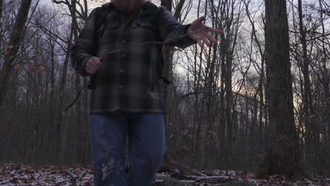 slow motion shot of a man with a big red beard wearing outdoor gear walking through the forest in winter breaking branches collecting fire wood for a campfire