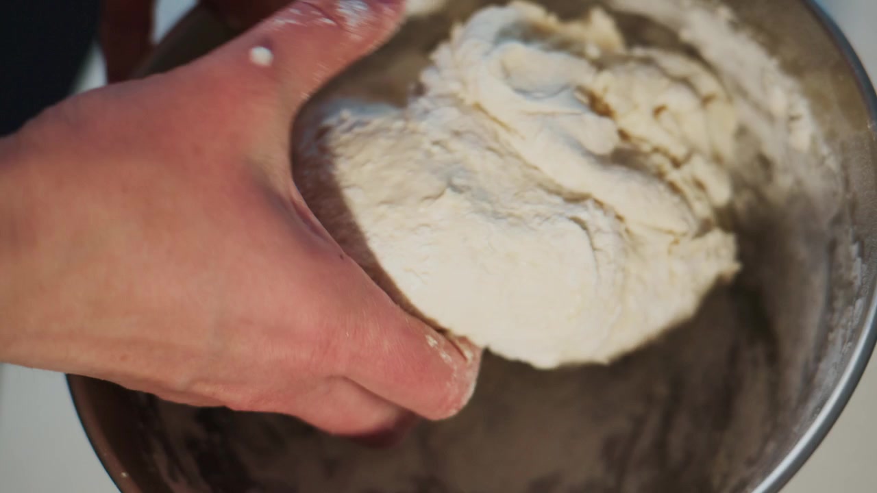 Hands knead dough in metal bowl. Flour dusts fingers and bowl surface. Person focused, calm, and skilled. Ideal for cooking, baking, or recipe videos. Shows traditional bread-making process