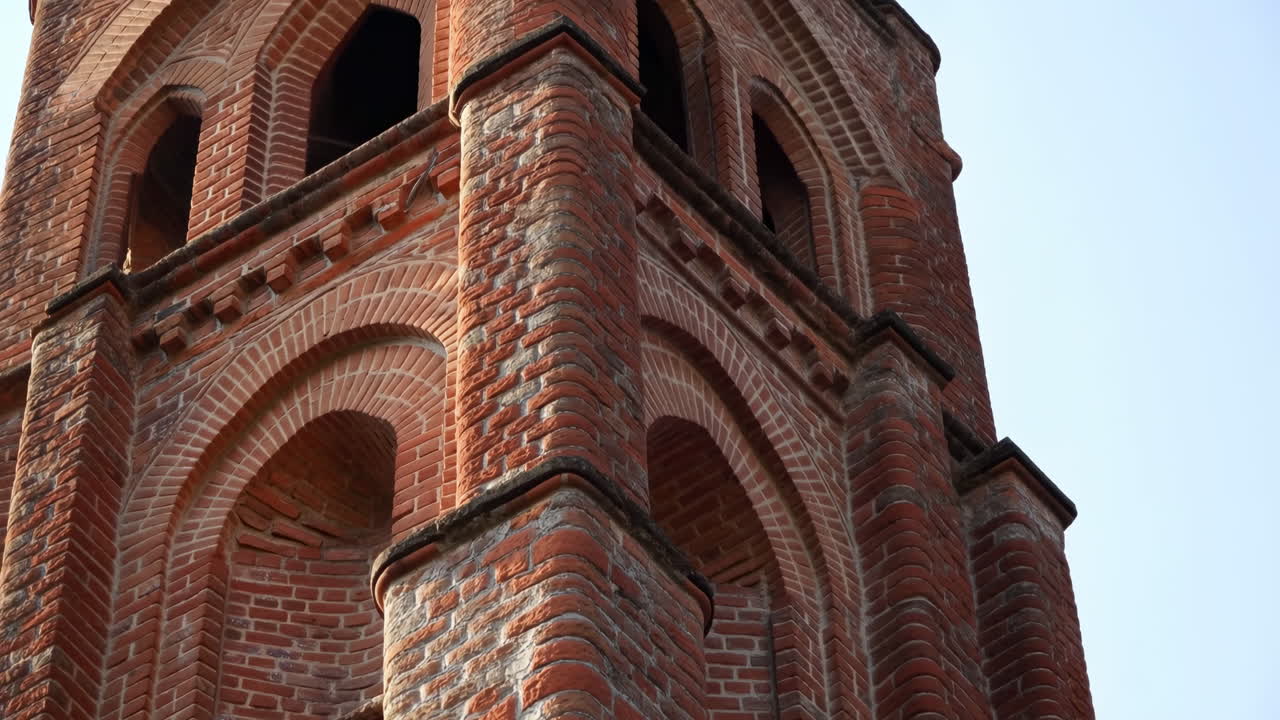 Detailní záběr červené cihlové zvonice, ukazující zvětralé zdivo, klenuté otvory, zdobenou římsu, vertikální perspektiva.