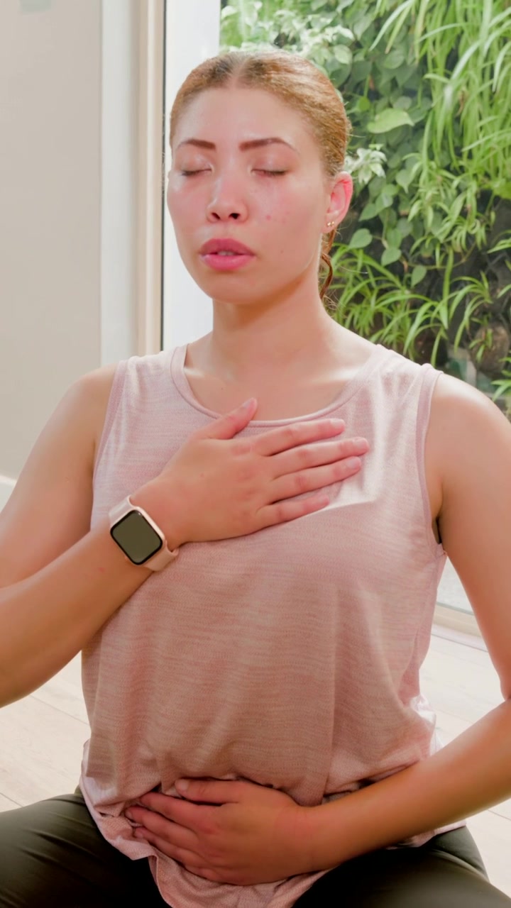 Video verticale: Donna in canotta rosa e leggings, ad occhi chiusi, che respira per rilassarsi vicino a una porta, con orologio.