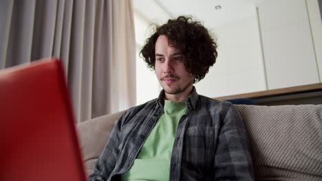 Confident-and-happy-brunette-guy-in-a-checkered-shirt-with-a-mustache-and-curly-hair-sits-on-the-sofa-and-works-on-a-red-laptop-at-home