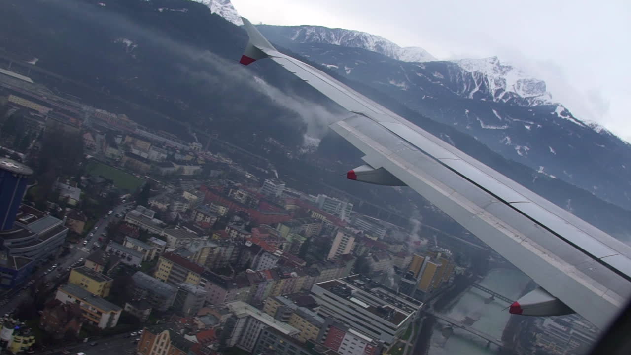Shot of airplane wing as plane comes in to land over the city of Innsbruck on a cloudy day, snowy mountains are in the background of the airport