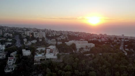 Ciudad-Con-Edificios-Y-Jardín-En-El-Borde-De-La-Costa-Al-Atardecer-Colorido,-Mar-De-Fondo,-Israel