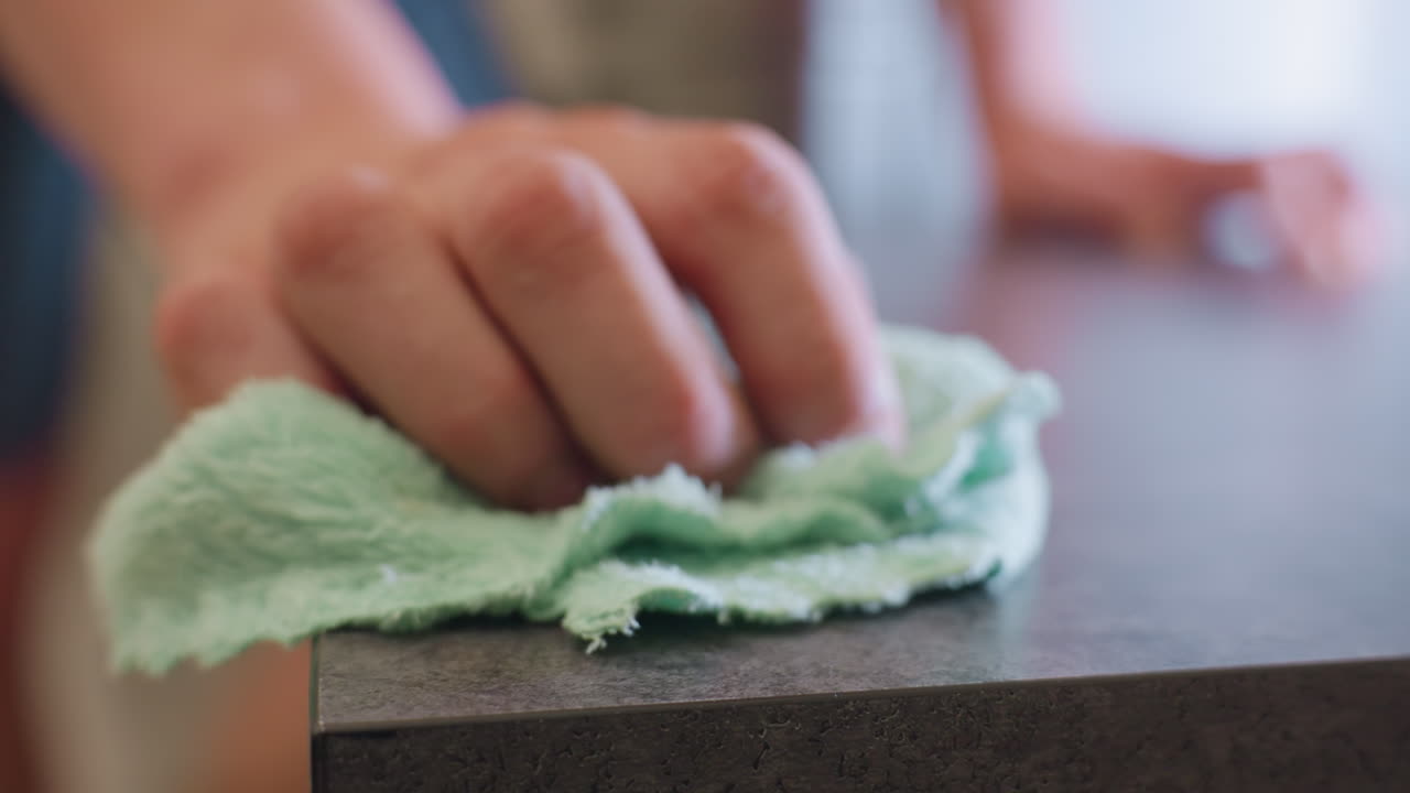 Close up partial view of hand cleaning wooden table using green cloth as sunlight reflects across smooth polished surface, showing motion forward for dust removal