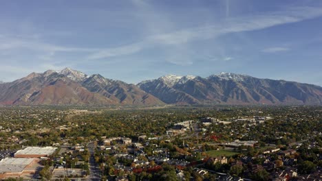 derecho camiones inclinar hacia abajo avión no tripulado amplio paisaje tiro de las impresionantes montañas rocosas cubiertas de nieve de utah con el condado de salt lake debajo lleno de edificios y árboles de colores en un cálido día soleado de otoño