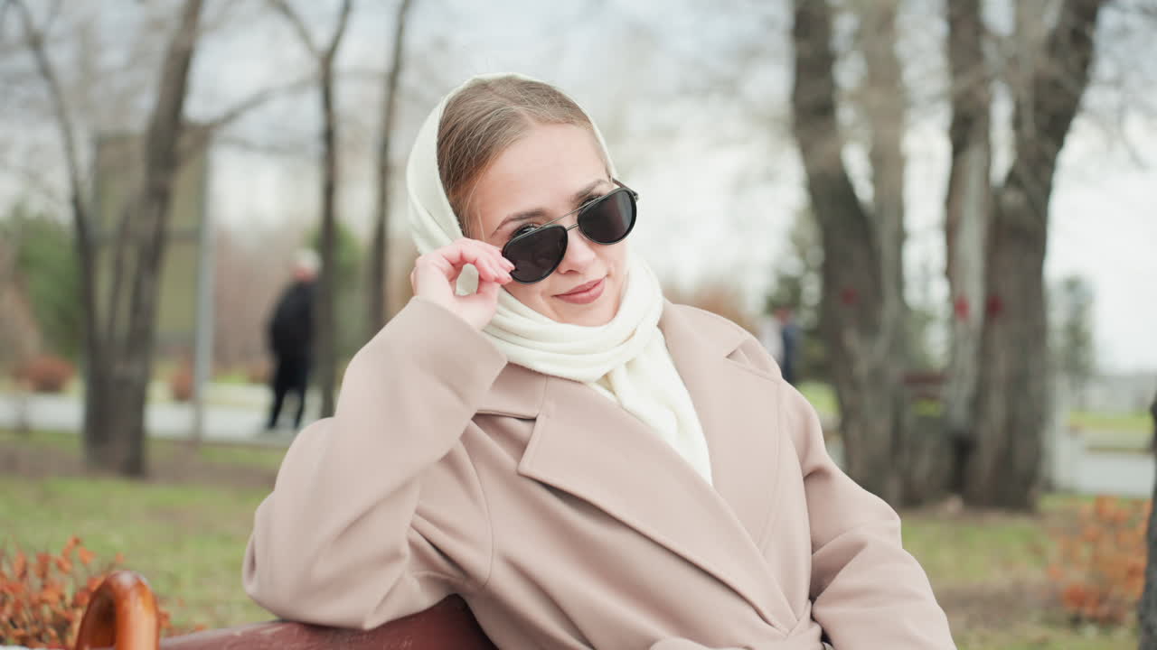 Elegant kaukasisk kvinde på en bænk i en stille efterårspark, iført beige frakke og hvidt tørklæde. Hun justerer solbrillerne, mens en vintage paraply hviler ved siden af hende. Hendes fremtoning udstråler en selvsikker modelvibe og en rolig, eftertænksom stemning.