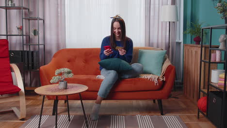 Woman-sitting-at-home-using-credit-bank-card-and-smartphone-while-transferring-money-online-shopping