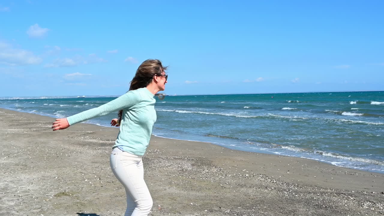 Cheerful woman in sunglasses and casual clothes jumping with joy on the sandy beach of Larnaca, Cyprus, with blue sea waves in the background