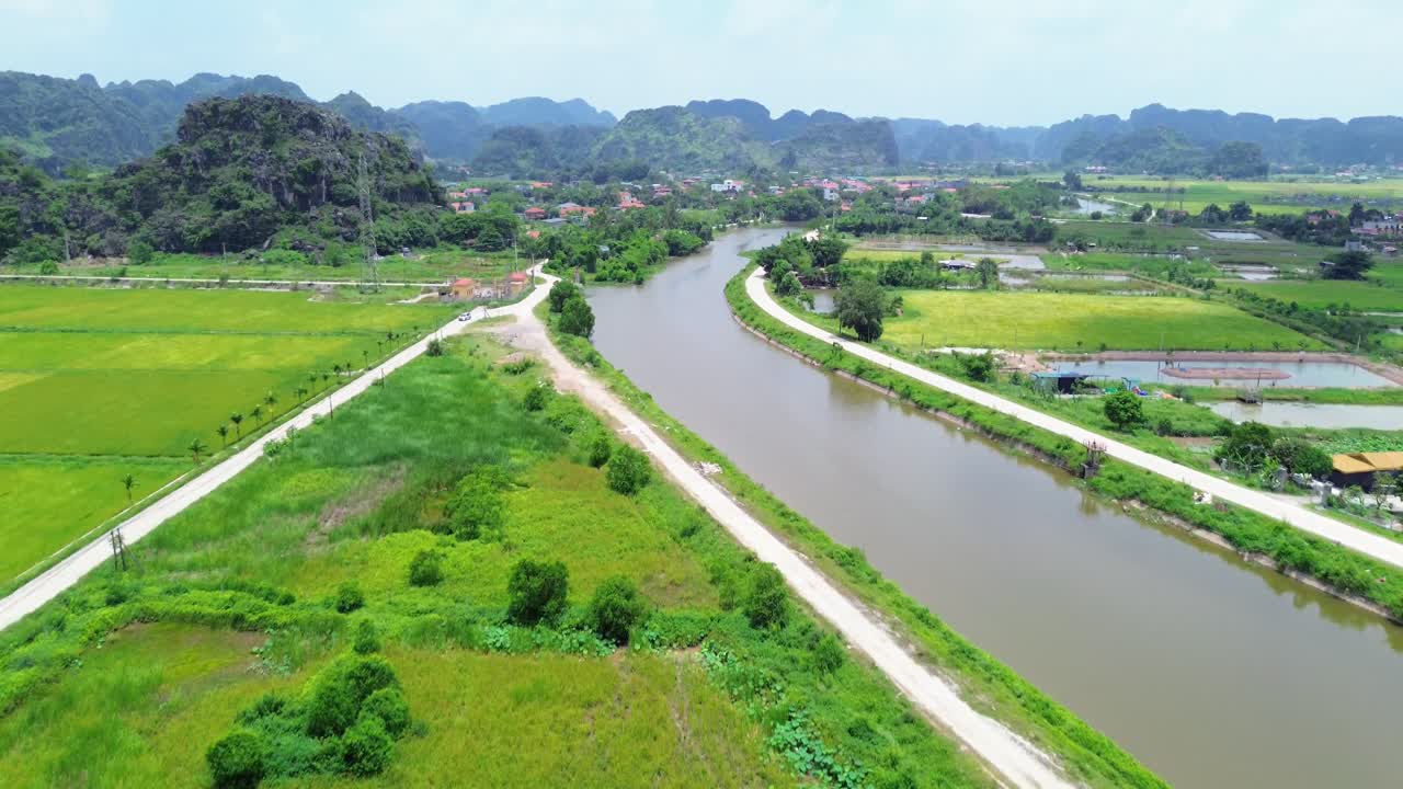 Aerial zoom in moving upward over winding river flanked by roads, rice paddies and distant karst hills