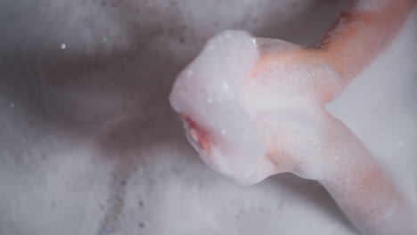 mujer juega con espuma esponjosa en la bañera de primer plano. dama sostiene un puñado de burbujas de jabón descansando en el baño acogedor. mujer se relaja durante la rutina higiénica