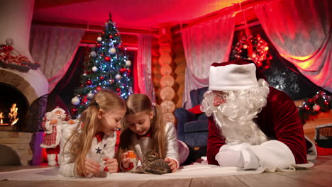 Happy children with kitten and Santa on floor. Little girls playing with a beautiful small cat near Santa Claus with white beard on the background of Christmas tree at home.