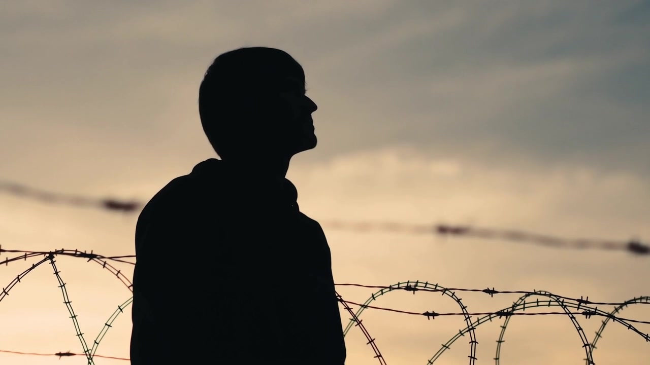 Standing man in hooded jacket tilting head near barbed coils, watching cloudy sky as dusk arriving