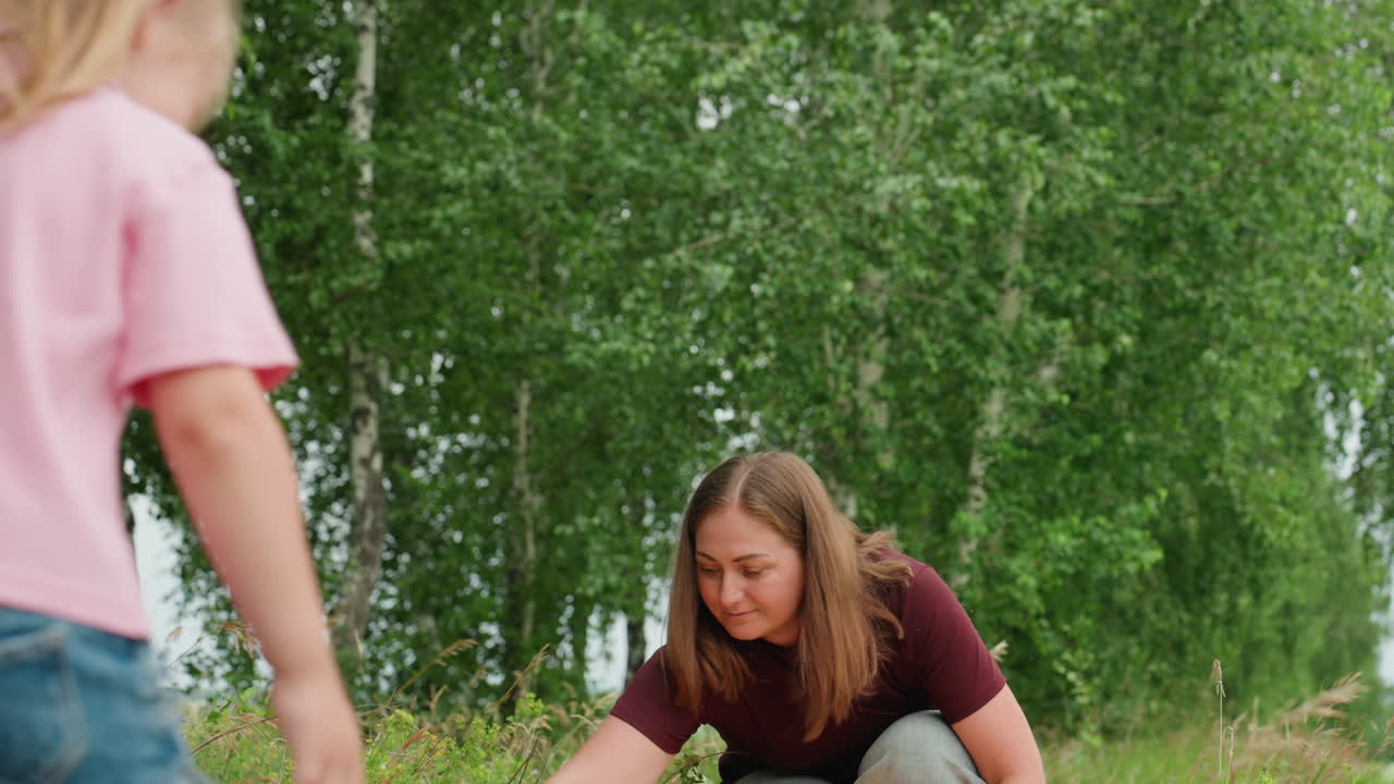 White woman teaching child in meadow adult kneels to examine ground while little girl leans forward, hands touching grass and soil, curious exploration, gentle guidance and focused learning