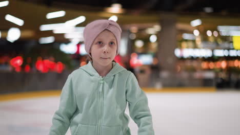 primer plano de una mujer con una capucha verde menta y una cinta rosa en la cabeza dirigiéndose a la cámara con una expresión enfocada, el fondo presenta luces borrosas y personas patinando en la pista de hielo