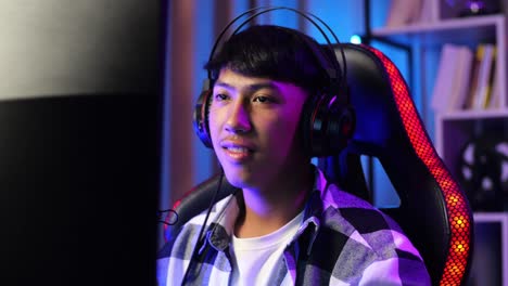 close up of asian boy streamer smiling and crossing his arms while playing game over network on personal computer. live stream video game, desk illuminated by rgb led strip light
